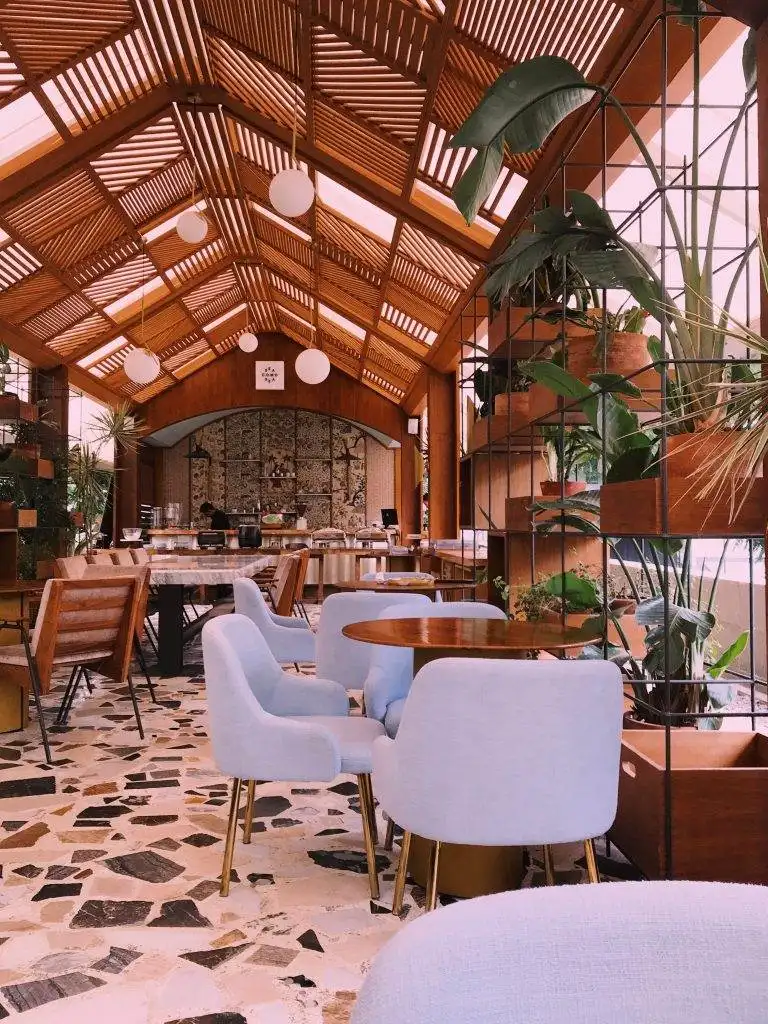 Modern restaurant interior with wooden ceiling, indoor plants, and pastel blue chairs creating a cozy dining atmosphere
